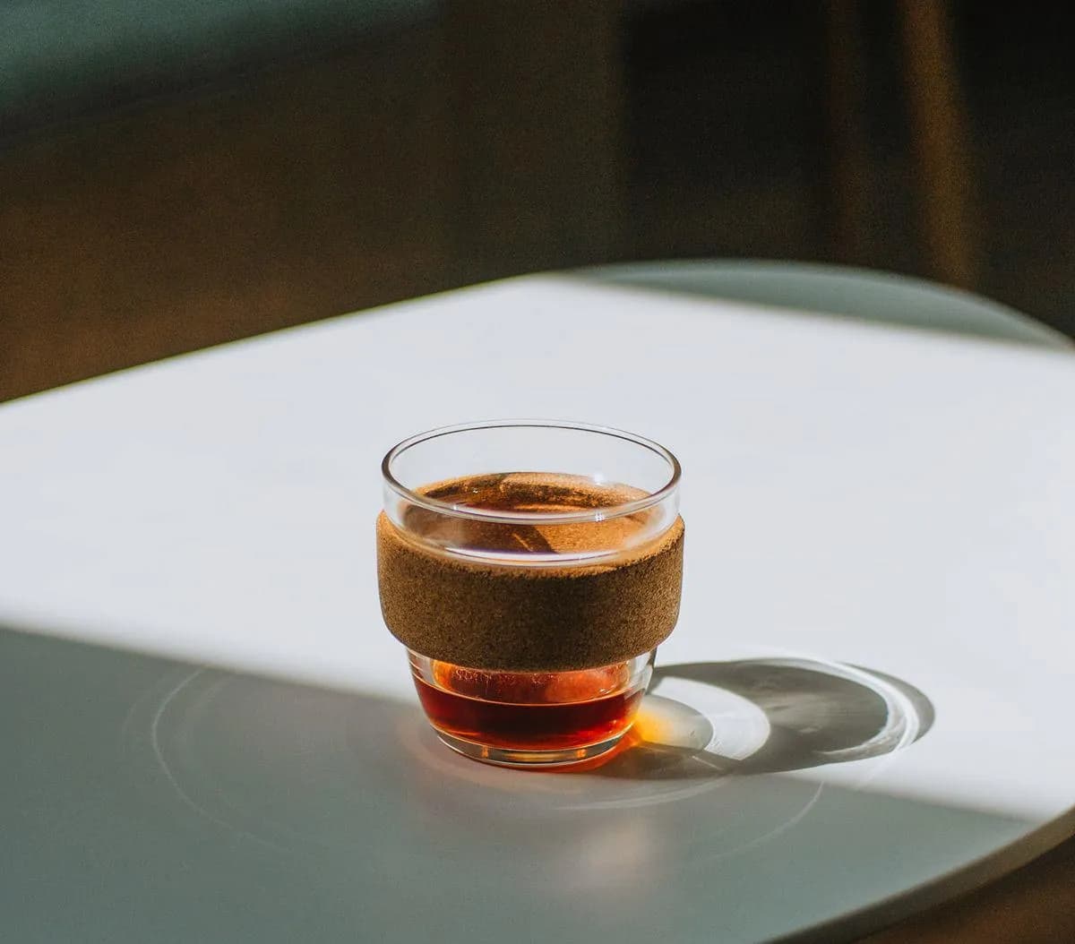 A glass cup of coffee with a cork sleeve on a sunlit table